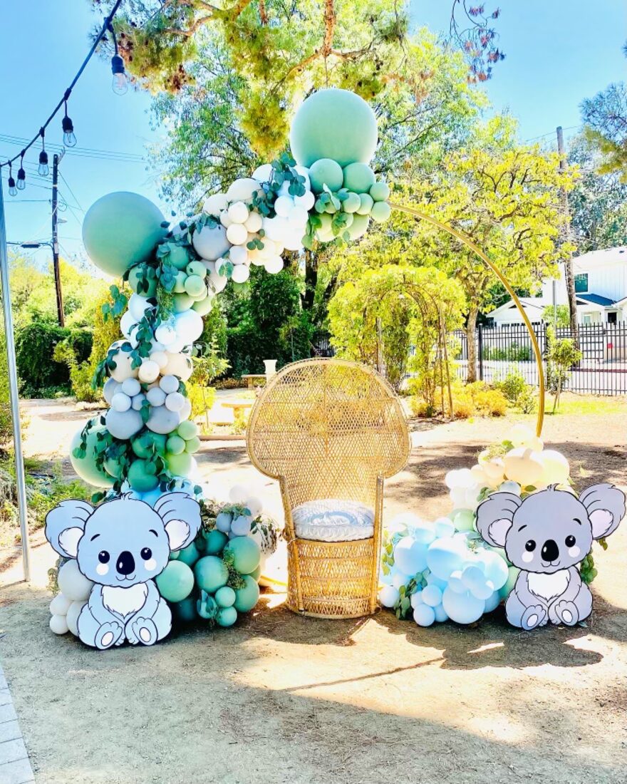 Sage and white balloon arch over a wicker peacock chair with koala cutouts